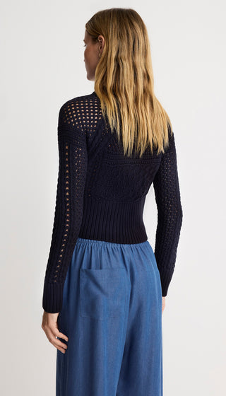 Woman wearing a black knit top with sheer sleeves and blue pants on a white background